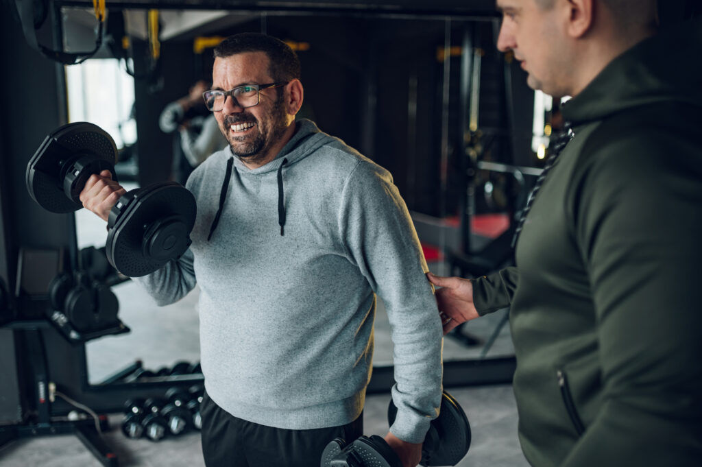 Middle aged men training in the gym with fitness trainer and using dumbbells.